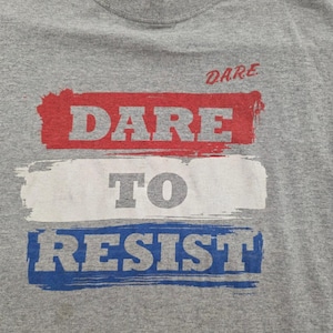 Puede incluir: Una camiseta gris con las palabras "DARE TO RESIST" en un diseño rojo, blanco y azul. La palabra "DARE" está encima de las otras palabras. La camiseta está hecha de un material suave.