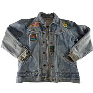May include: Light blue denim jacket with multiple patches. Patches include an American flag, a bear with the text "Thank You", and the text "I'm in a good space". The jacket has a button-up front, two chest pockets, and two side pockets.