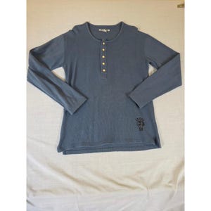 Op de afbeelding: Een blauw Henley-shirt met lange mouwen, een knoopsluiting en een ronde hals. Het shirt heeft een kleine afbeelding aan de onderkant rechts en is gemaakt van een zachte, getextureerde stof. De knopen zijn geel.