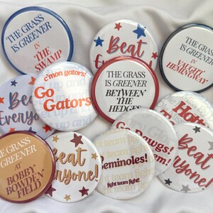 May include: A collection of round buttons with various phrases and designs. Some buttons feature text like "The Grass is Greener" and "Beat Everyone!" in red, white, and blue. Others say "Go Gators" and "Seminoles!" with star accents.