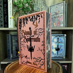Op de afbeelding: Een hardcover boek getiteld "Shield of Sparrows" met een roze kaft en zwarte en gouden illustraties. Het boek heeft een zwaard- en vogelontwerp. Andere boeken zijn zichtbaar op een houten plank.