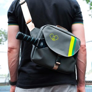Könnte beinhalten: Graue Umhängetasche mit gelb-grünem Streifen und gelbem Pickleball-Logo. Ein schwarzer und türkisfarbener Pickleball-Schläger befindet sich in der Seitentasche. Die Tasche hat einen beigen Riemen und eine Schnalle.