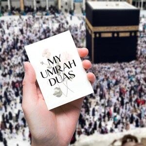 May include: A white card with the text "MY UMRAH DUAS" in black lettering and floral illustrations. The card is held in a hand with a blurred background of a large crowd of people in white clothing gathered around a black cube.