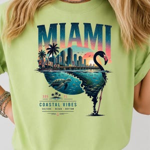 May include: A light green t-shirt featuring a graphic print of the Miami skyline, a sunset, a sea turtle, and a flamingo silhouette. The text reads MIAMI, 305, FL, COASTAL VIBES, CULTURE, OCEAN, RHYTHM. The design incorporates blue, orange, and pink colors.