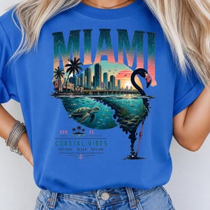 May include: A bright blue t-shirt featuring a graphic print of the Miami skyline, a sea turtle, and a flamingo silhouette. The design includes the text MIAMI, 305 FL, COASTAL VIBES, CULTURE, OCEAN, RHYTHM, and a small anchor icon in a sunset color palette.