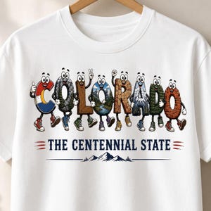 Puede incluir: Una camiseta blanca con la palabra COLORADO deletreada por personajes de dibujos animados, donde cada letra representa un paisaje o textura diferente. Debajo, el texto dice THE CENTENNIAL STATE junto a un gráfico de montañas.