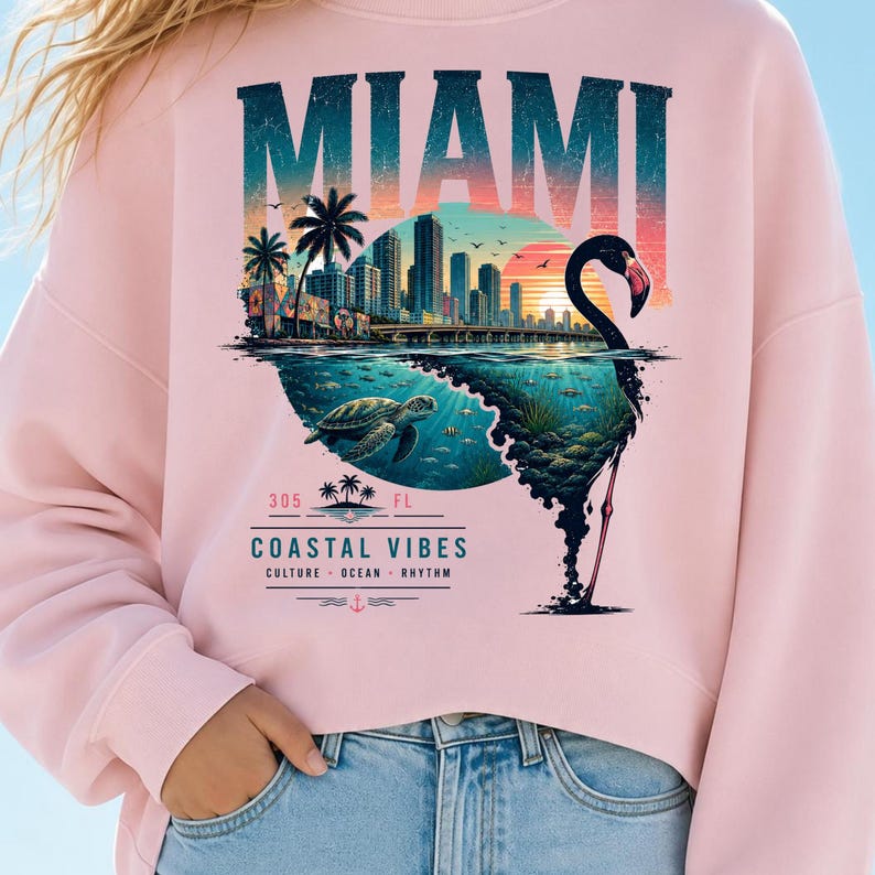 May include: A light pink sweatshirt featuring a graphic print of the Miami skyline, a sea turtle, and a flamingo silhouette. The text reads MIAMI, 305 FL, COASTAL VIBES, CULTURE, OCEAN, RHYTHM. The design incorporates sunset colors, palm trees, and water.