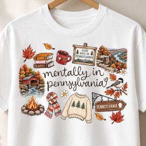 May include: A white t-shirt featuring a graphic print with the text "mentally in pennsylvania" surrounded by illustrations of books, a coffee mug, a covered bridge, a campfire, a scarf, a sweater, a bird, autumn leaves, and signs.