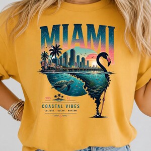 May include: A mustard yellow t-shirt featuring a graphic print of the Miami skyline at sunset, a sea turtle swimming underwater, and a flamingo silhouette. The text reads MIAMI, 305 FL, COASTAL VIBES, CULTURE, OCEAN, RHYTHM.