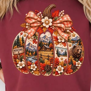 May include: A maroon t-shirt featuring a large pumpkin graphic filled with a mountain landscape, a log cabin, a bear, and a sign that reads MONTANA with a heart. The pumpkin is decorated with a plaid bow, white flowers, berries, and pinecones.
