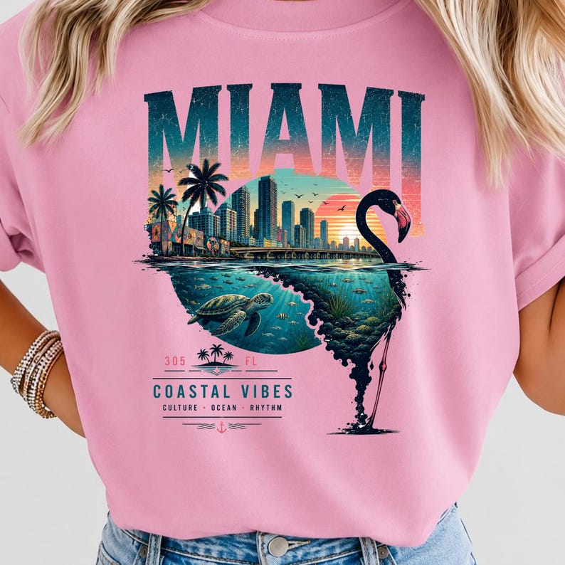 May include: A light pink t-shirt featuring a graphic print of the Miami skyline at sunset, a sea turtle swimming underwater, and a flamingo silhouette. The text reads MIAMI, 305, FL, and COASTAL VIBES CULTURE OCEAN RHYTHM in a stylized font.