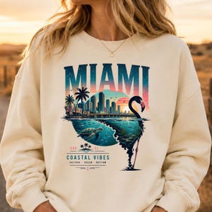 May include: A cream-colored crewneck sweatshirt featuring a graphic print of the Miami skyline, a sea turtle, and a flamingo silhouette. The text reads MIAMI, 305, FL, COASTAL VIBES, CULTURE, OCEAN, RHYTHM, with a small anchor icon at the bottom.