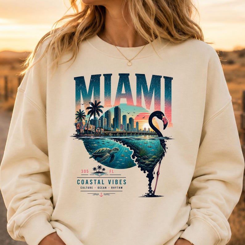 May include: A cream-colored crewneck sweatshirt featuring a graphic print of the Miami skyline, a sea turtle, and a flamingo silhouette. The text reads MIAMI, 305, FL, COASTAL VIBES, CULTURE, OCEAN, RHYTHM, with a small anchor icon at the bottom.