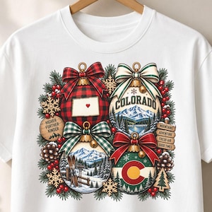 May include: A white t-shirt featuring a graphic of four Christmas ornaments surrounded by pine branches, red berries, and snowflakes. The ornaments display a red plaid pattern, a Colorado mountain landscape, a snowy ski scene, and the Colorado state flag.