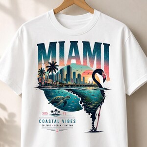 May include: A white t-shirt featuring a graphic print with the text MIAMI at the top. The design includes a city skyline, a sunset, a sea turtle swimming underwater, and a flamingo silhouette. Below, the text 305 FL COASTAL VIBES CULTURE OCEAN RHYTHM.