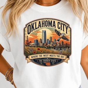 May include: A white t-shirt featuring a graphic of the Oklahoma City skyline at sunset. The design includes a flying scissor-tailed flycatcher, an oil derrick, sunflowers, and the text 'OKLAHOMA CITY WHERE THE WEST MEETS THE SKY THE SOONER STATE' with a map of Oklahoma.