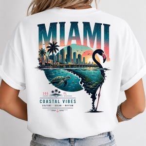 May include: A white t-shirt featuring a graphic on the back with the text MIAMI in large letters above a city skyline, a sunset, a sea turtle swimming underwater, and a flamingo silhouette. Below is the text 305 FL COASTAL VIBES CULTURE OCEAN RHYTHM.