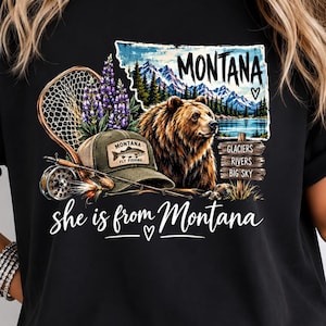 May include: A black t-shirt featuring a graphic of a brown bear, a fishing net, a cap with a fish logo, and a mountain landscape. The text reads MONTANA, GLACIERS, RIVERS, BIG SKY, and she is from Montana with a heart symbol included in the design.