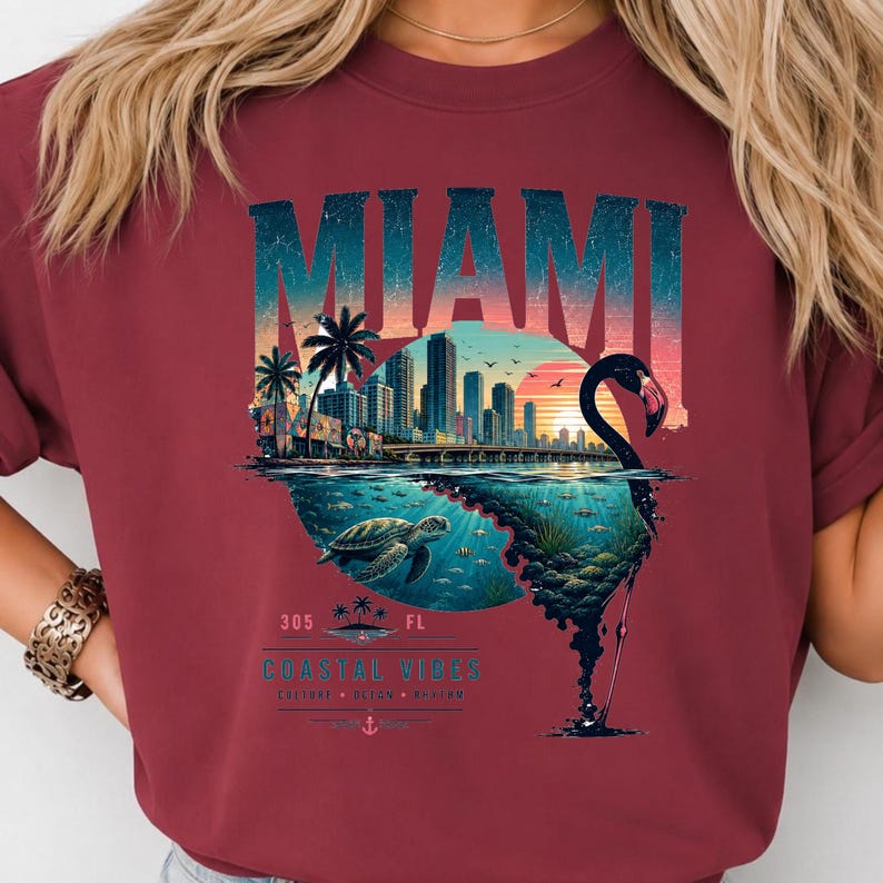 May include: A maroon t-shirt featuring a graphic of the Miami skyline at sunset, a sea turtle swimming underwater, and a flamingo silhouette. The text reads MIAMI, 305 FL, and COASTAL VIBES. The design incorporates palm trees and an anchor icon.