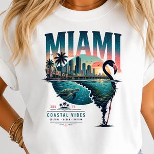 May include: A white t-shirt featuring a graphic print of the Miami skyline at sunset, a sea turtle swimming underwater, and a flamingo silhouette. The text reads MIAMI, 305 FL, COASTAL VIBES, CULTURE, OCEAN, RHYTHM, with palm tree and anchor icons.