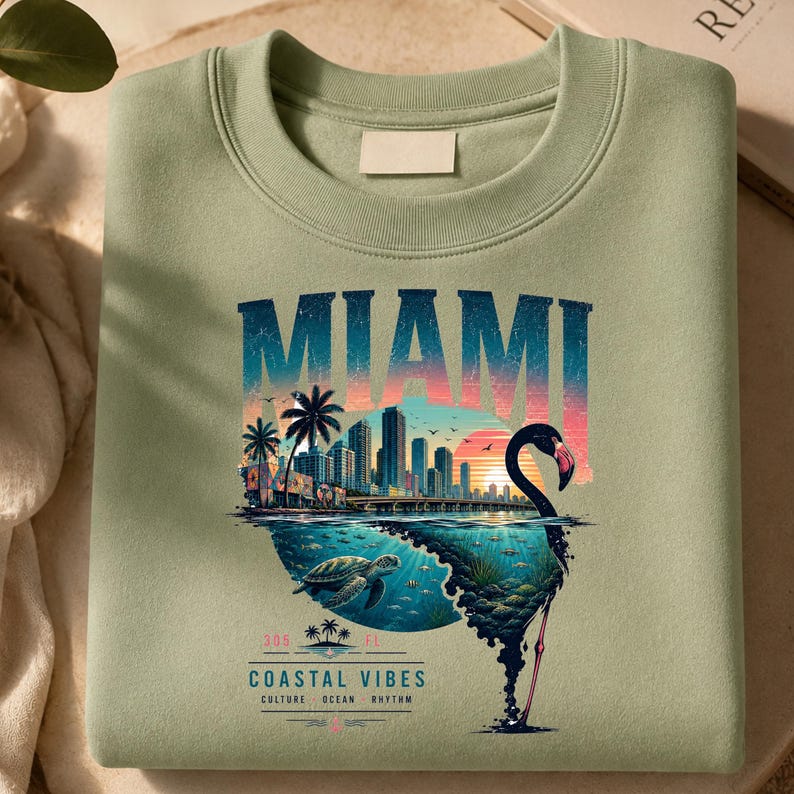 May include: A sage green crewneck sweatshirt featuring a graphic print of the Miami skyline at sunset. The design includes a flamingo silhouette, a sea turtle, palm trees, and the text MIAMI, 305 FL, COASTAL VIBES, CULTURE, OCEAN, RHYTHM.