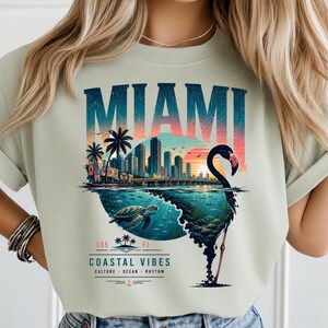 May include: A light green t-shirt featuring a graphic print of the Miami skyline at sunset, a sea turtle swimming underwater, and a large flamingo silhouette. The text reads MIAMI, 305 FL, COASTAL VIBES, CULTURE, OCEAN, RHYTHM.