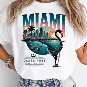 May include: A white t-shirt featuring a graphic print of the Miami skyline at sunset, a sea turtle swimming underwater, and a flamingo silhouette. The text reads MIAMI, 305 FL, COASTAL VIBES, CULTURE, OCEAN, RHYTHM, with palm trees and an anchor icon.