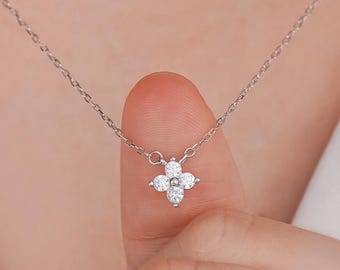 May include: A delicate silver necklace featuring a four-petal flower pendant. The pendant is adorned with small, sparkling white gemstones and a central pink gemstone. The necklace is displayed against a white shirt.