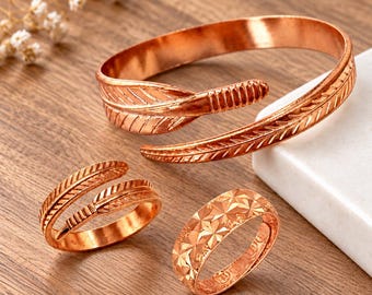 May include: A copper jewelry set consisting of an open cuff bracelet with a feather design, an open ring with a matching feather motif, and a band ring featuring a repeating geometric star pattern across the entire outer surface.