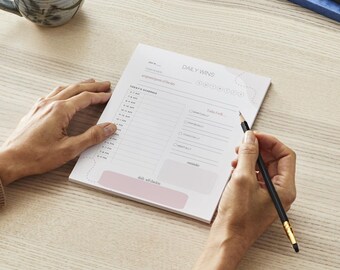 Könnte beinhalten: Ein weißer Planer mit einem rosa Abschnitt am unteren Rand. Der Planer hat einen Tagesplan, einen Abschnitt für tägliche Erfolge und einen Abschnitt für die heutigen Ziele. Ein Bleistift liegt auf dem Planer.