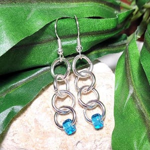 May include: Silver-toned dangle earrings with a chain-like design. Each earring features multiple connected rings and a small, square, turquoise-colored bead at the bottom. The earrings are displayed against a backdrop of green leaves.