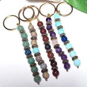 May include: A collection of beaded keychains with gold-colored rings. Each keychain features a different color scheme of gemstone beads, including green, brown, purple, and blue. The beads are separated by decorative metal spacers.