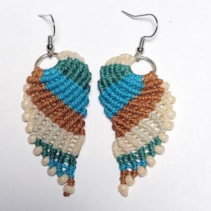 May include: A pair of handmade macrame earrings. The earrings feature a wing-like design with alternating stripes of teal, blue, brown, and cream-colored cord. Small, off-white beads are attached to the bottom edge. Silver-colored ear wires complete the design.