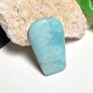 May include: A polished, light blue gemstone with a smooth, rounded shape. The stone has subtle variations in color and texture, with a slightly translucent appearance. The gemstone is set against a neutral background.
