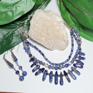 May include: A layered necklace and matching earrings set. The necklace features multiple strands of blue and silver beads, with elongated blue stone pendants. The earrings have a similar design, with blue beads and silver cone-shaped accents.
