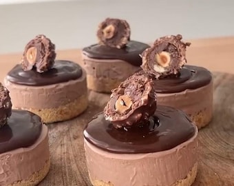 May include: Five individual chocolate cheesecakes topped with chocolate ganache and a whole or half Nutella chocolate hazelnut spread candy on a wooden cutting board.