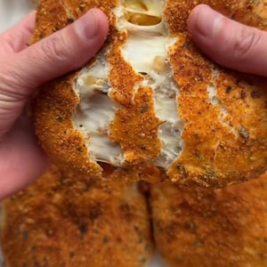 Può includere: Primo piano di una mano che tiene un panino rotondo dorato con ripieno di formaggio. Il pane è ricoperto da un condimento rosso e verde. Il pane è diviso, rivelando il formaggio filante all'interno. Il pane è su una superficie bianca con altri panini simili.
