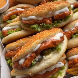May include: A close-up of a stack of small sandwiches with chicken, lettuce, pickles, and a creamy sauce. The sandwiches are on a white paper liner in a light brown tray.