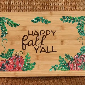 May include: A rectangular bamboo cutting board with the words "HAPPY FALL Y'ALL" in brown script. The board is decorated with green leaves and red pumpkins.