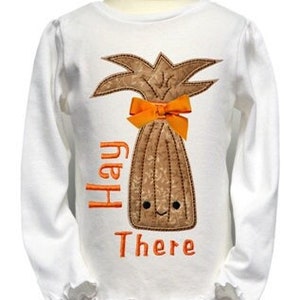 May include: White long-sleeved shirt with an embroidered brown hay bale with a smiling face and an orange bow. The text "Hay There" is embroidered below the bale.