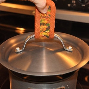 Puede incluir: Una olla de plata con tapa y asa. La tapa tiene un asa de plata con un agarrador de olla de tela marrón y naranja con estampado.