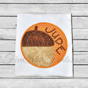 May include: A round orange and brown applique with a brown acorn design and the name "Jude" in white text.