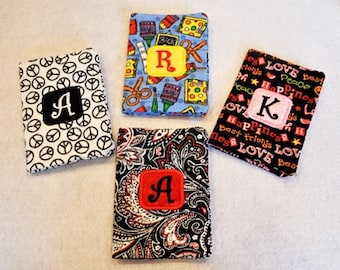 May include: Four fabric covered notebooks with personalized embroidered letters. The notebooks have different patterns, including peace signs, paisley, school supplies, and words like "love" and "happiness".