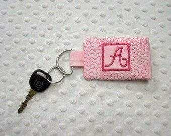 May include: Pink fabric key chain with a pink embroidered letter A in a square frame. The key chain is attached to a silver key ring.