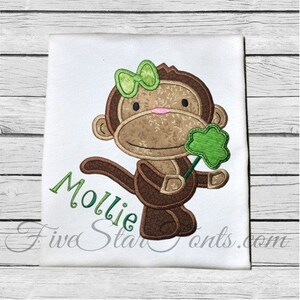 Pode incluir: Desenho bordado de um macaco marrom com um laço verde e segurando um trevo verde. O nome "Mollie" está bordado abaixo do macaco.