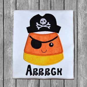 May include: A white t-shirt featuring an orange and yellow candy corn wearing a black pirate hat with a skull and crossbones. The text "ARRGH" is below the candy corn.