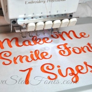 Puede incluir: Una máquina de bordar está cosiendo el texto "Make Me Smile Font 7 Sizes" en hilo naranja sobre una tela blanca. El texto está en una fuente cursiva.