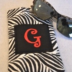 May include: A black and white zebra print sunglasses case with a red embroidered initial "GJ" on a black square patch.