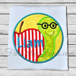 May include: A green worm with glasses is peeking out from behind a red and white striped apple. The word "Liam" is embroidered on the apple. The design is in a circle with a blue border.