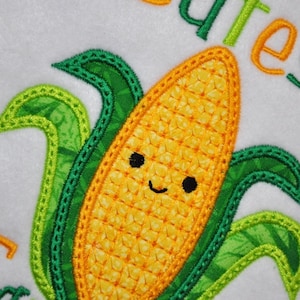 May include: Embroidered design of a smiling corn cob with green leaves. The corn is yellow with a textured pattern, and the leaves are green with a patterned fabric. The word "cute" is embroidered in green and orange.